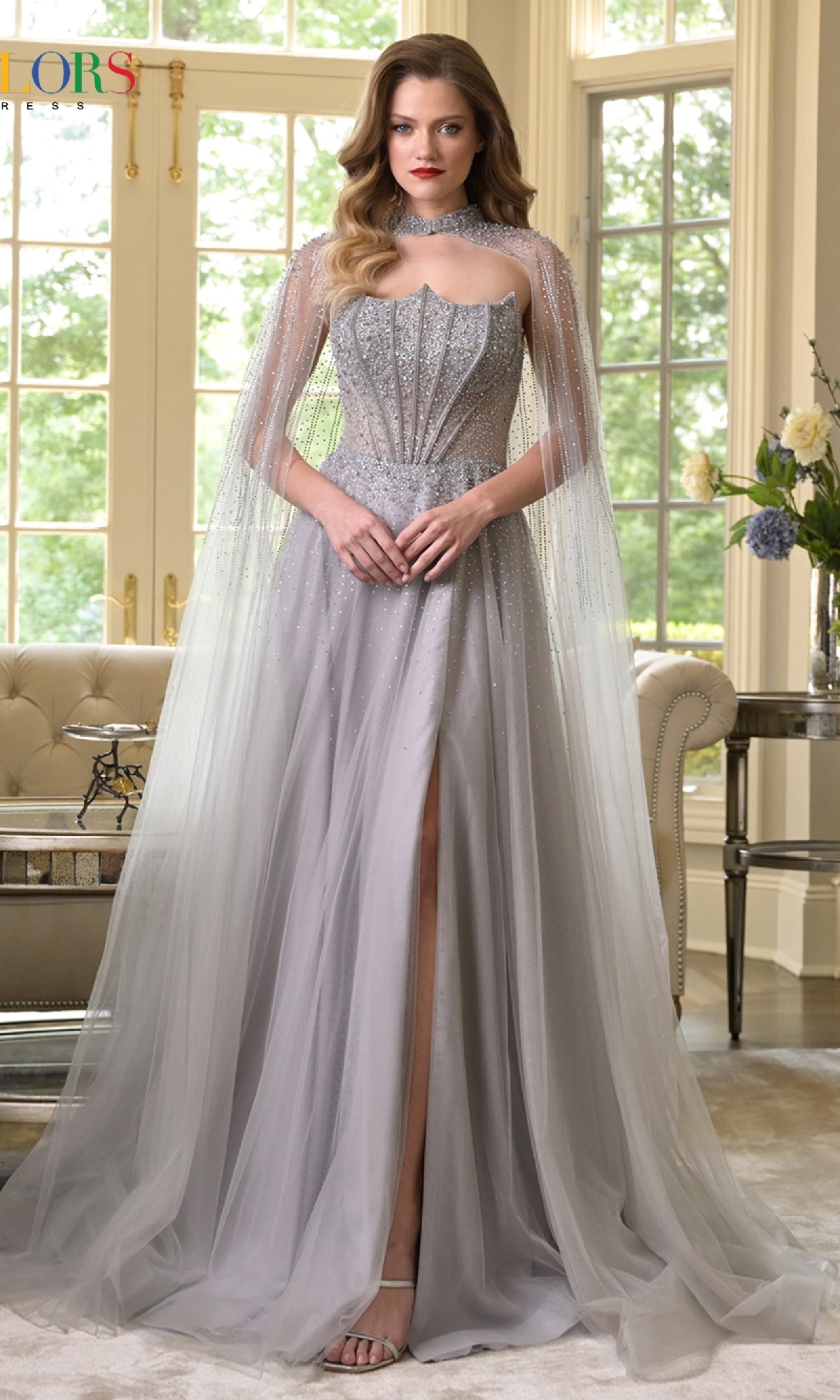 A woman poses indoors in the Colors Dress 3879, a silver, floor-length prom gown with an embellished corset bodice, sheer cape, and thigh-high slit. Large windows behind her showcase lush greenery outside.